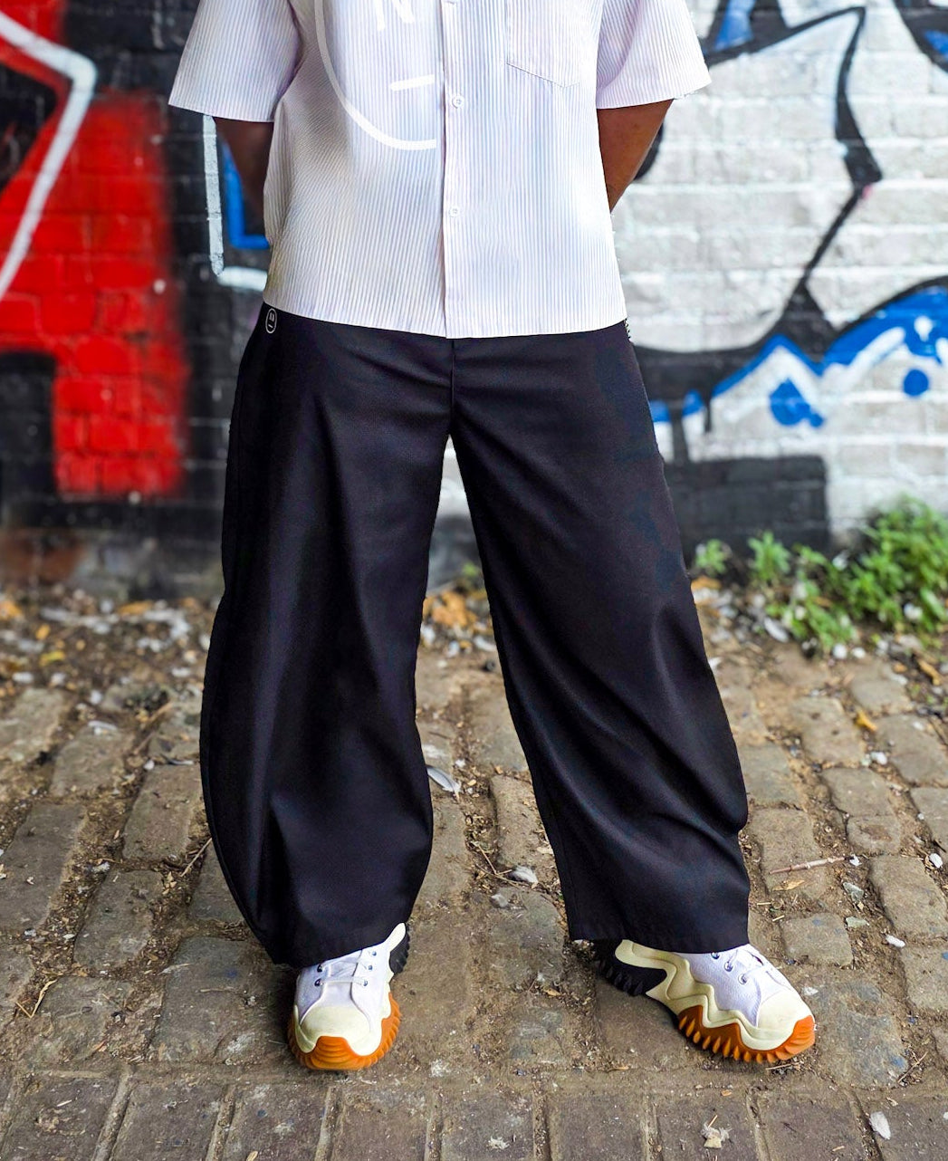 Man wearing a white shirt and black pants standing in front of a graffiti wall.