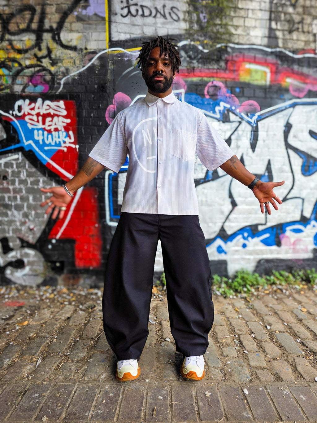 Person wearing a white shirt with a logo and dark pants standing in front of graffiti art.