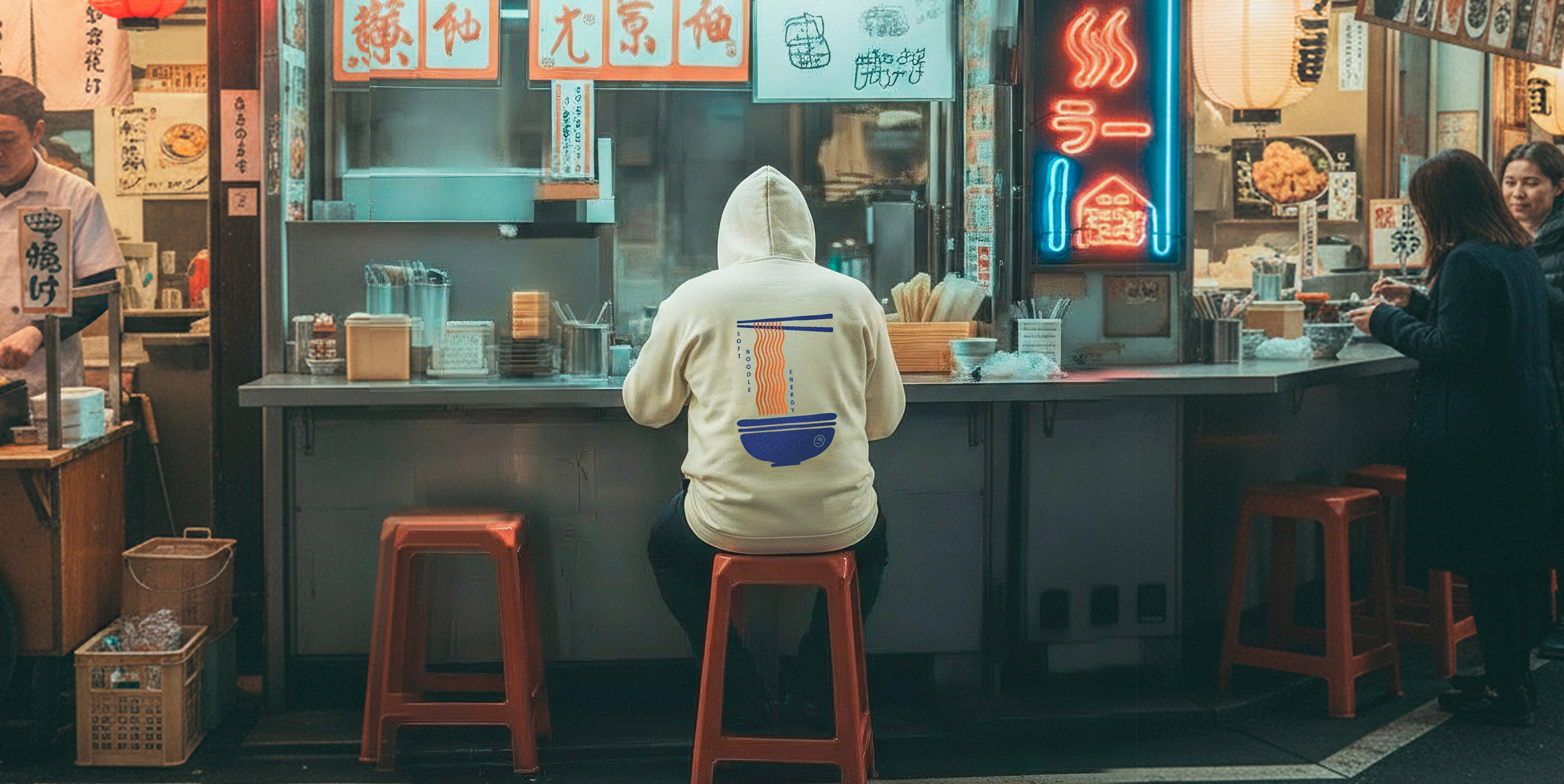 Person sitting at a counter in a busy street food market with neon signs and people around.