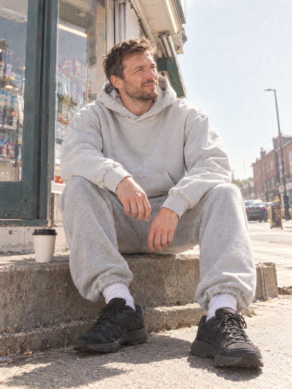 Man wearing the Snow Wash Oversized Hoodie in Light Gray with grey sweatpants, sitting on a shop step
